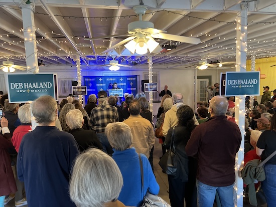 A group of people stands in a room listening to Deb Haaland