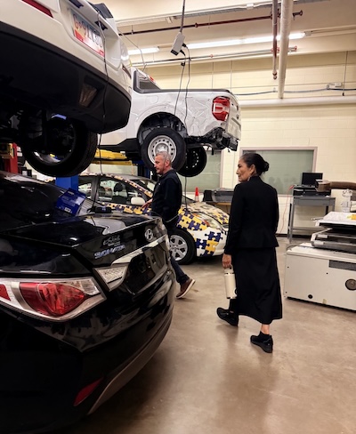 Deb Haaland in a garage, observing a car elevated on a platform above them.